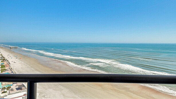 On peut admirer la plage depuis une fenêtre, avec un ciel dégagé, une plage de sable et une jetée qui se dessine au loin.