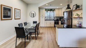 Une cuisine moderne avec des meubles blancs, un îlot central doté d’un plan de travail noir, et un coin repas avec une table et des chaises en verre.