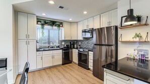 Une cuisine moderne avec des meubles blancs, des appareils en acier inoxydable, un plan de travail noir et une fenêtre avec des stores.