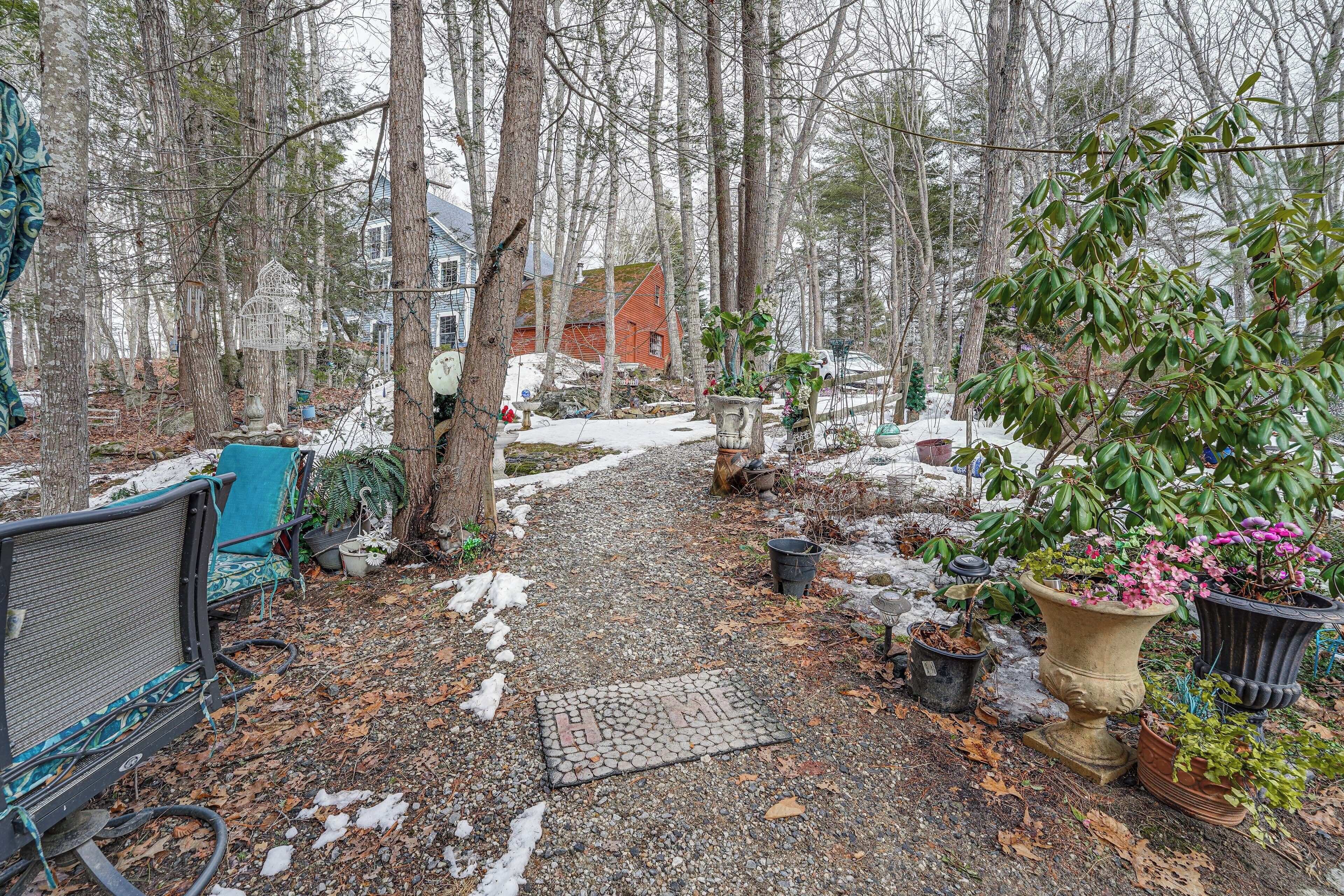 A gravel path leading to a house surrounded by trees, with potted plants and a bench.