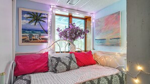 Ein Bett mit gemusterten Betttextilien, eine Vase mit Blumen und Wandbilder, darunter eine Strandansicht und ein Sonnenuntergang.