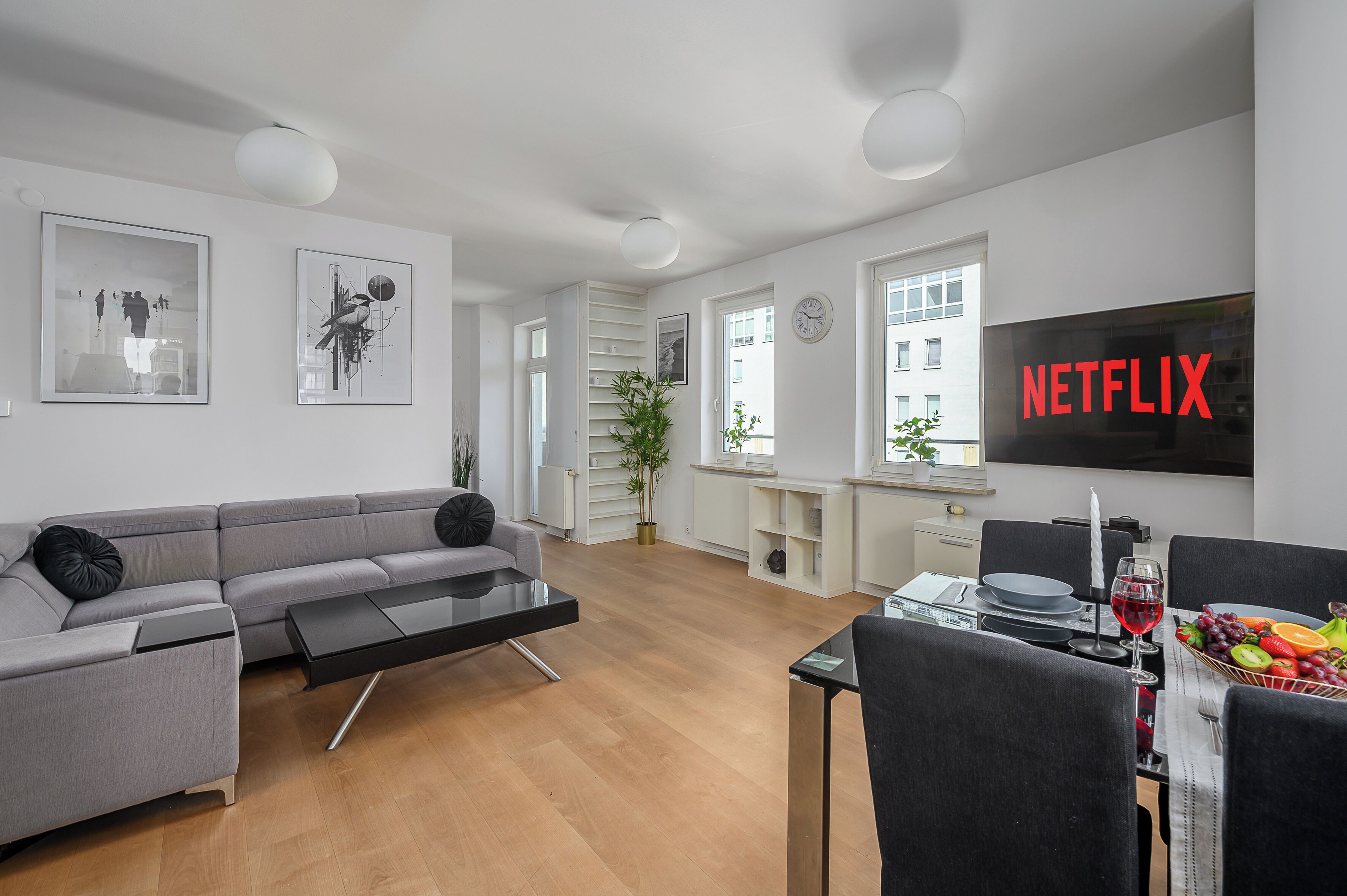 Una sala de estar moderna con un sofá gris, una mesa de comedor con fruta y vino, y un televisor que muestra 'NETFLIX'.