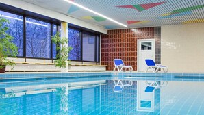 En indendørs swimmingpool med krystalklart vand, to liggestole og udsigt til træer gennem store vinduer.