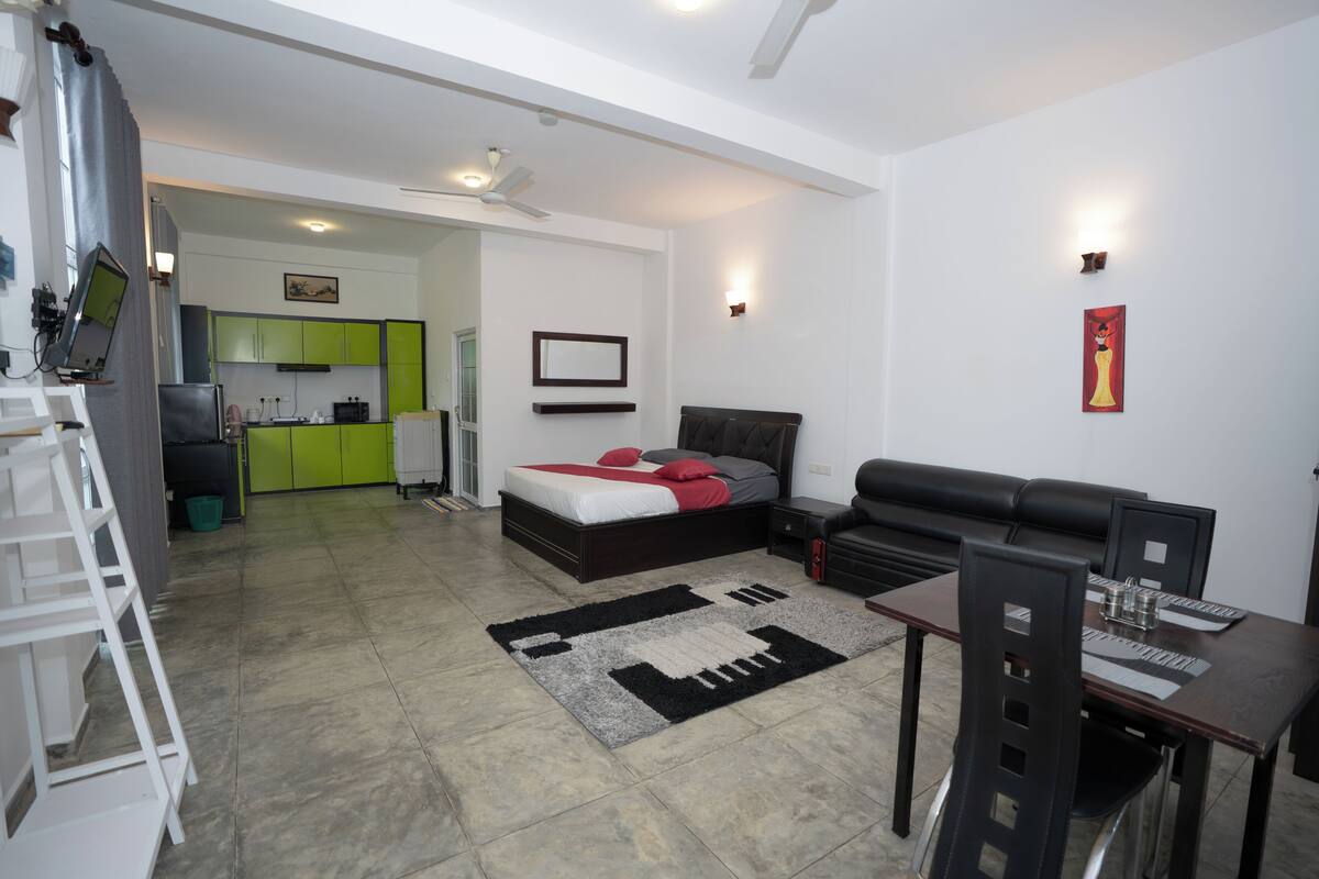 A modern living room with a bed, a dining table, and a kitchen in the background.