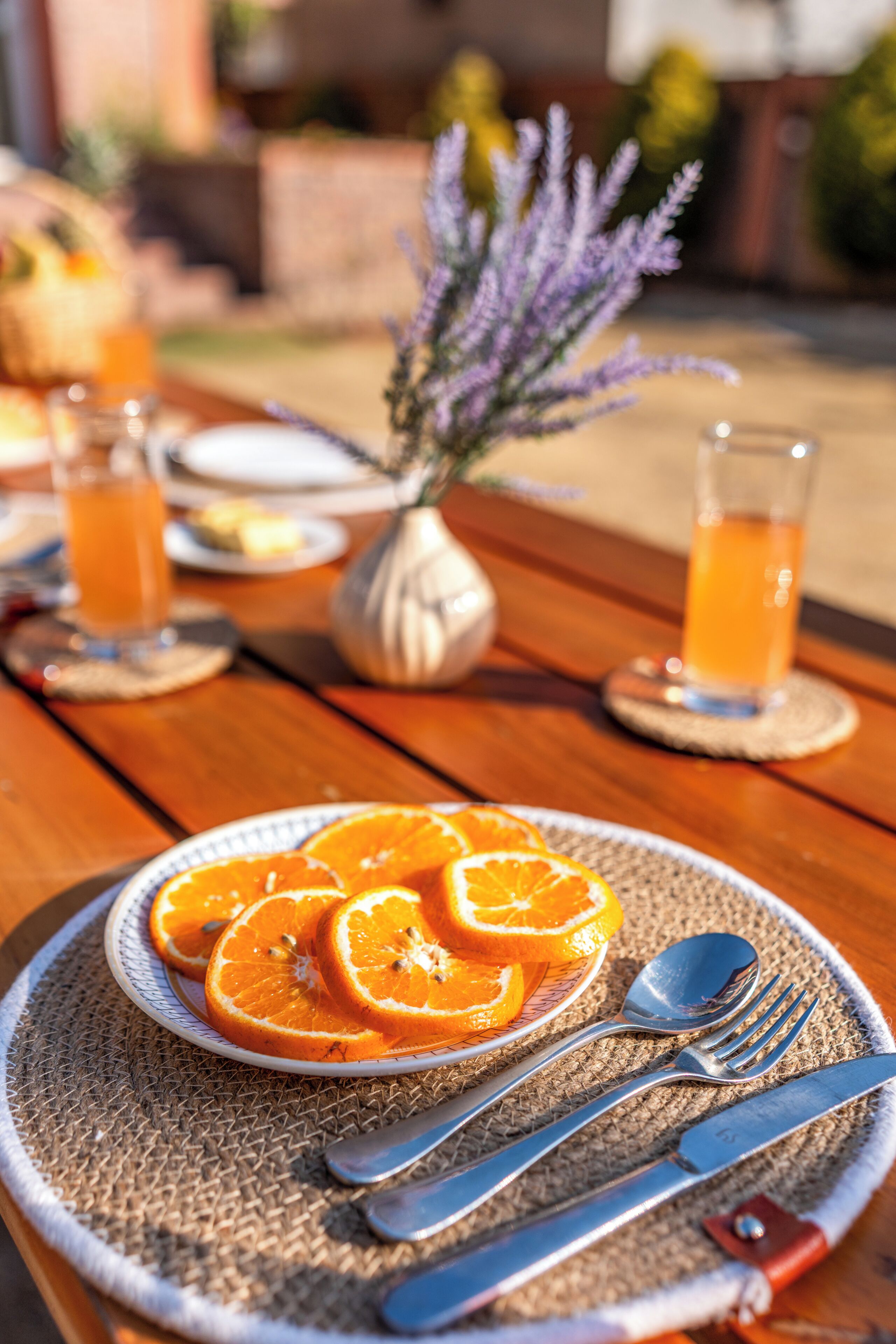A wooden table with a plate of orange slices, a vase with purple flowers, and a glass of orange juice.