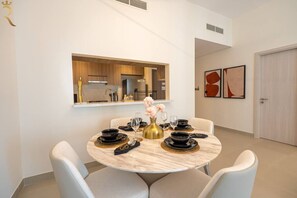 A dining area with a round table, white chairs, a kitchen in the background, and a framed artwork on the wall.