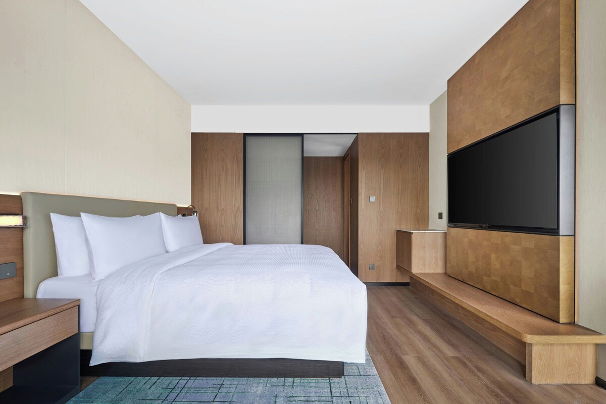 A modern hotel room with a large bed, a flat-screen TV mounted on the wall, and a wooden wardrobe.