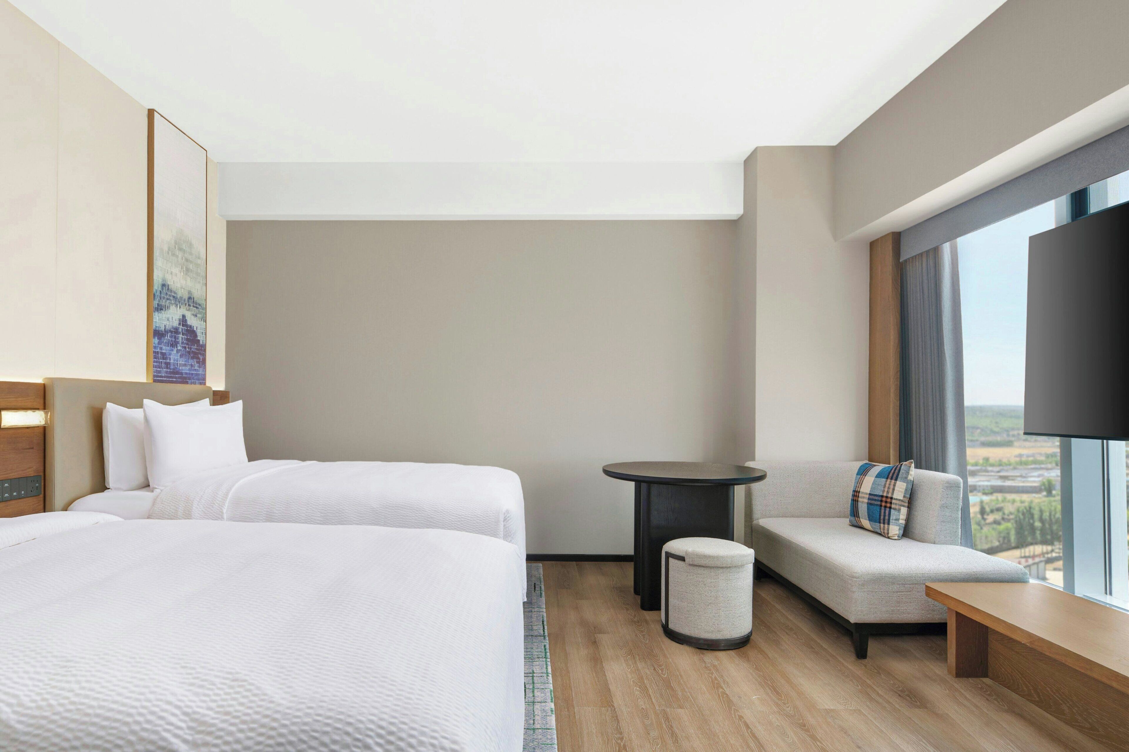 A hotel room with two beds, a sofa, a small round table, a TV, and a large window with a view.