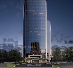 Un hotel moderno de gran altura con fachada de vidrio, entrada bien iluminada y un área ajardinada con árboles.