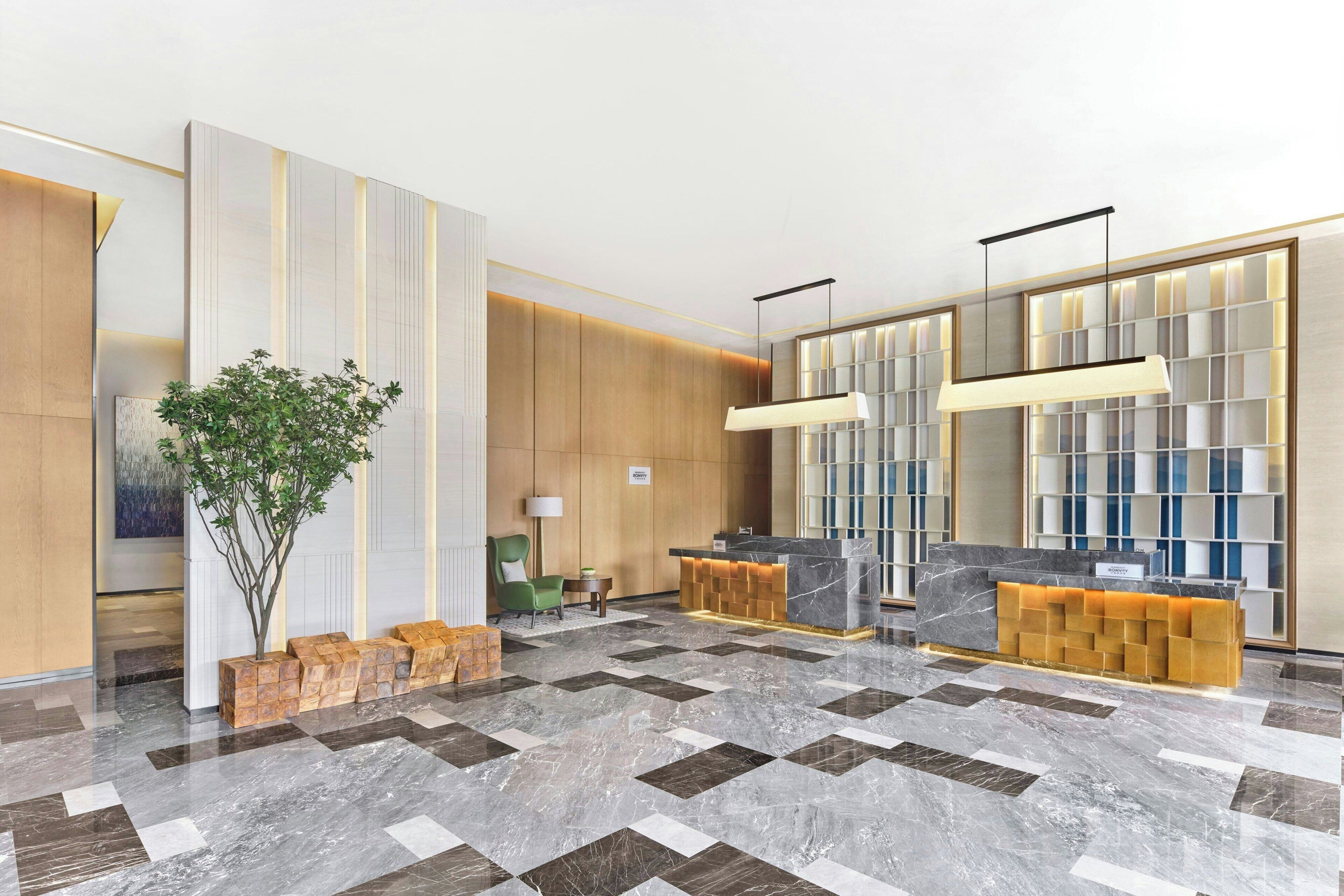 A modern hotel lobby with a reception desk, a green chair, a plant, and a decorative wall with a grid pattern.