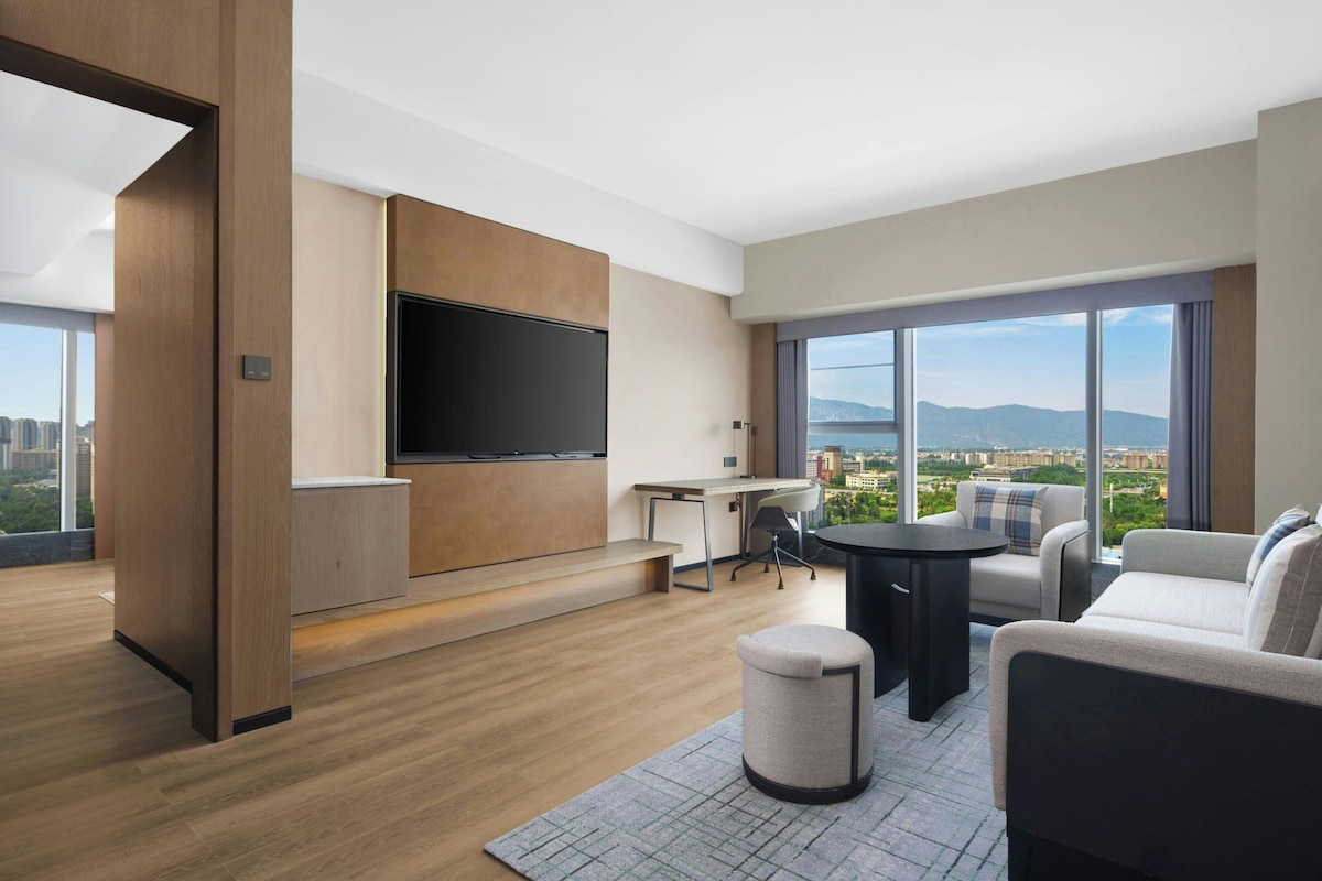 A modern hotel room with a large TV, a desk, a sofa, and a view of the city.