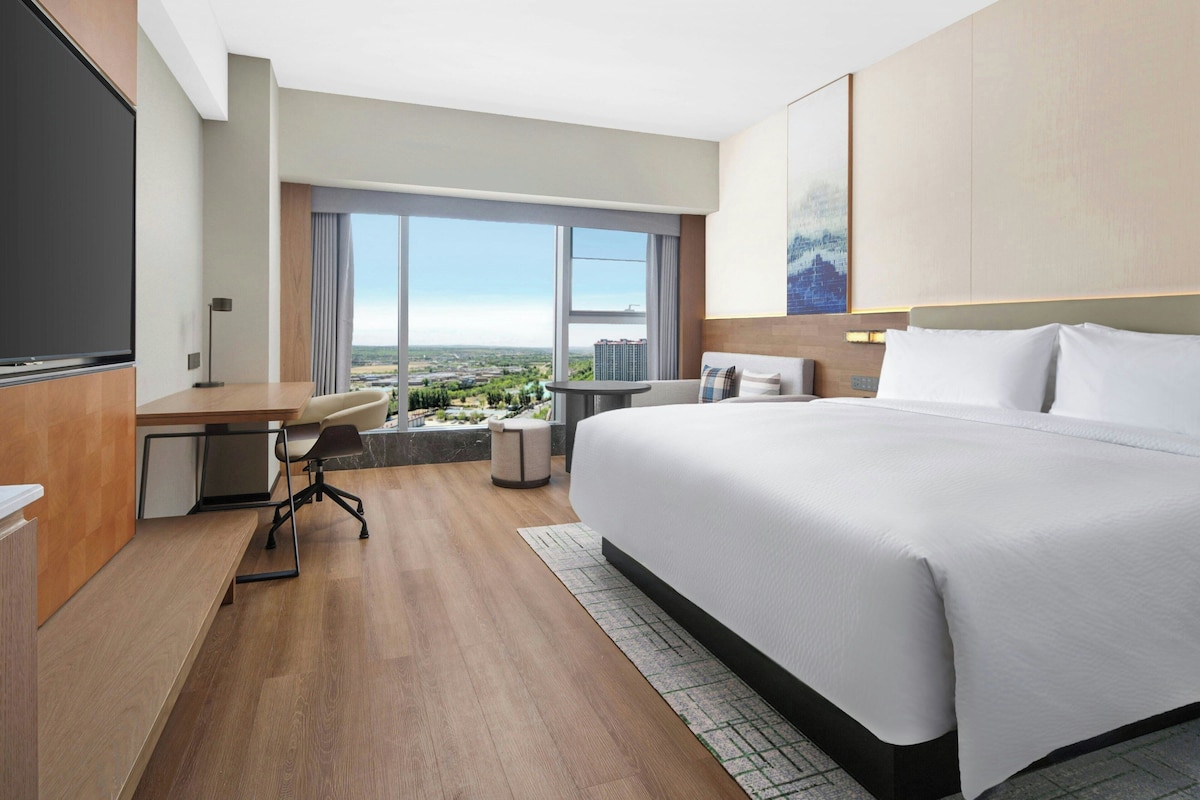 A hotel room with a large bed, a desk, a chair, and a view of the cityscape through the window.