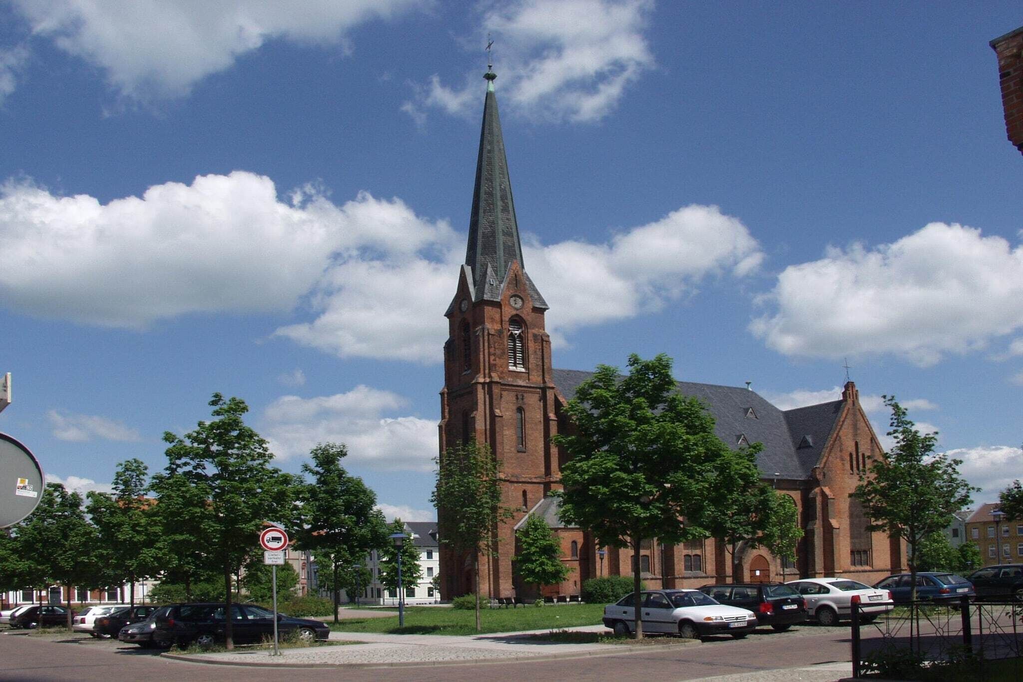 Una chiesa con un campanile alto, circondata da alberi e auto parcheggiate.