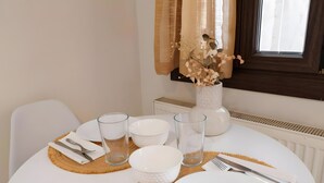 Une table à manger dressée avec de l’assiette blanche, des bols, des verres et des couverts, et un vase de fleurs séchées posé sur le rebord de la fenêtre.