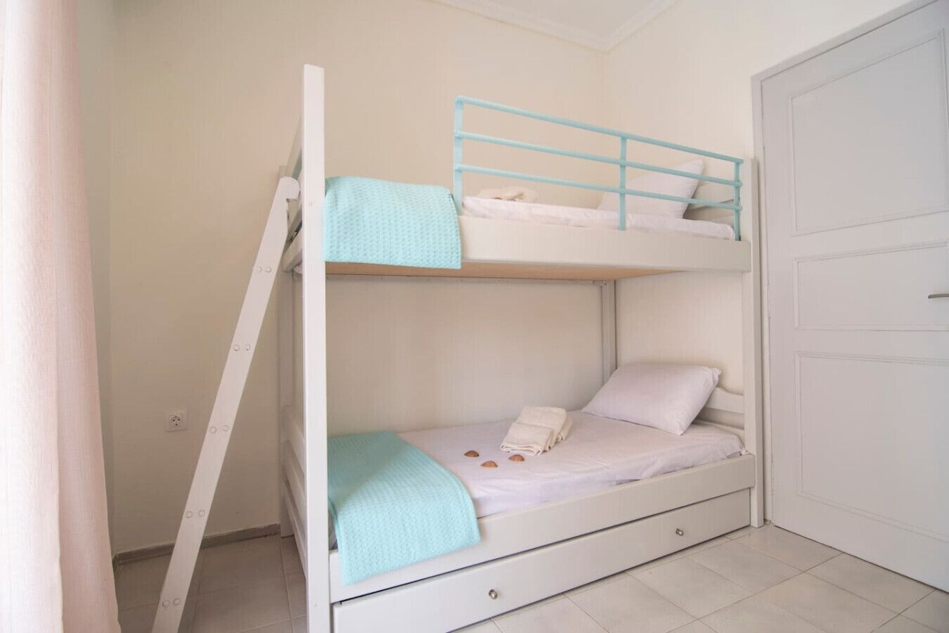 A bunk bed with a ladder, a drawer, and a door in a room with white walls and a tiled floor.