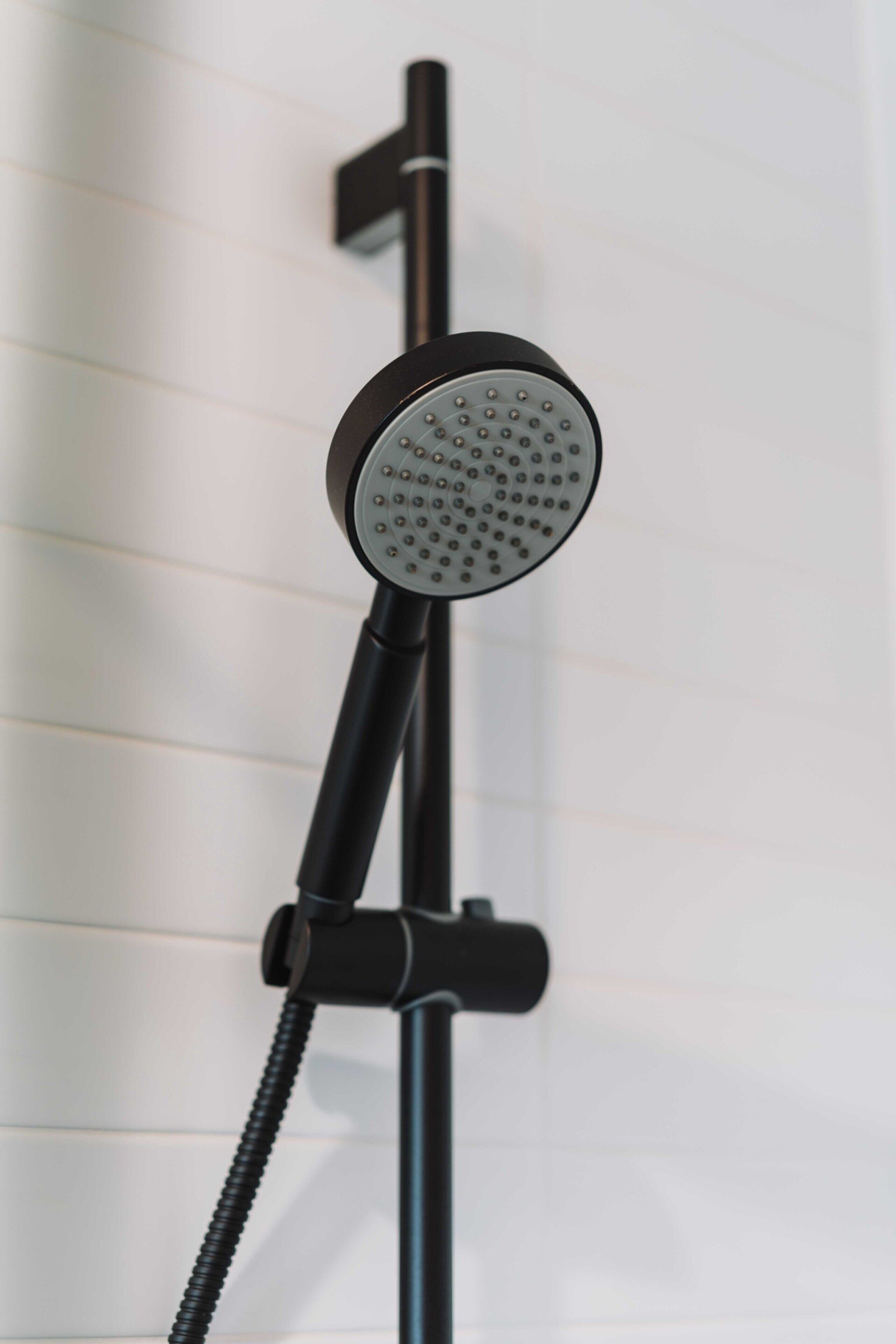 Une pomme de douche moderne, dotée d’un flexible, fixée sur un support noir devant un mur carrelé.