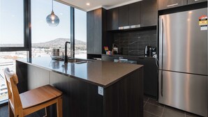 A modern kitchen with a large island, dark cabinetry, stainless steel appliances, and a view of a cityscape. - Lovely Sunny 2-bedroom Apartment With Pool and Gym (Canberra)