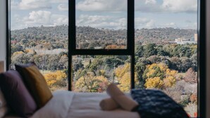 A bed with pillows, a window with a view of a hilly landscape and buildings. - Lovely Sunny 2-bedroom Apartment With Pool and Gym (Canberra)