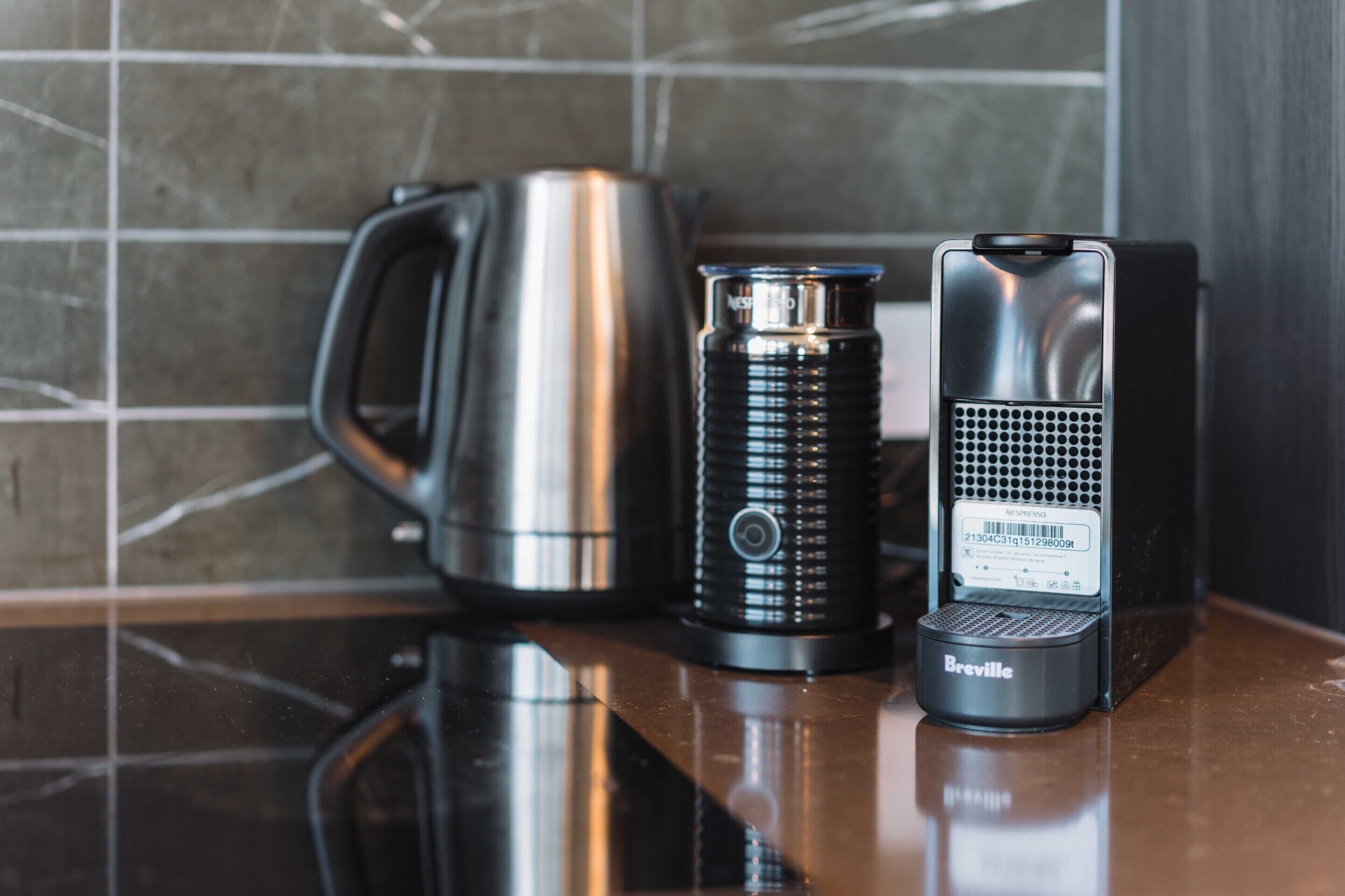 Eine Breville Kaffeemaschine, ein schwarzer Wasserkocher und ein Edelstahl-Kaffeekännchen auf einer spiegelnden Arbeitsplatte.