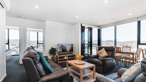 A modern living room with leather sofas, a wooden coffee table, and a TV. Large windows offer a city view. - Lovely Sunny 2-bedroom Apartment With Pool and Gym (Canberra)