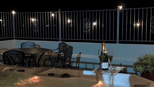 Un bain à remous avec une table et des chaises, entouré d’une rambarde et illuminé de guirlandes lumineuses.