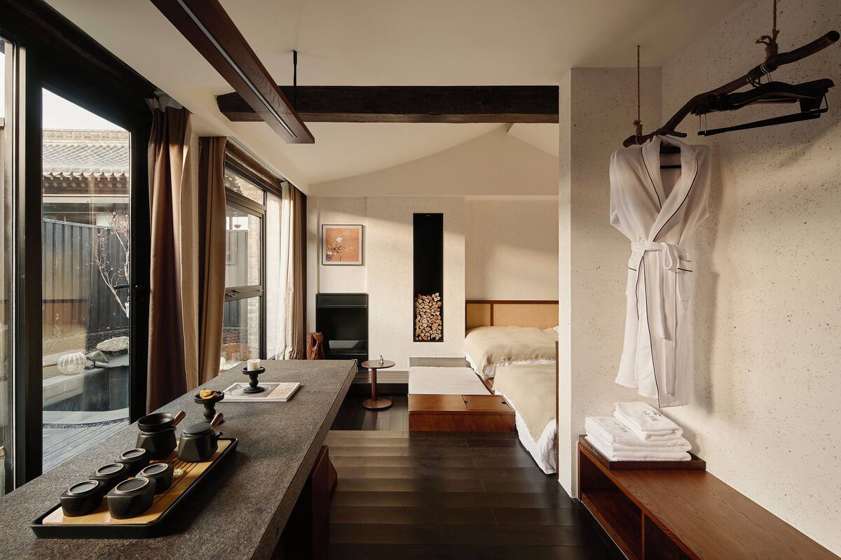 A hotel room with a wooden floor, a bed, a TV, a small table with a tray of tea cups, and a window with curtains.