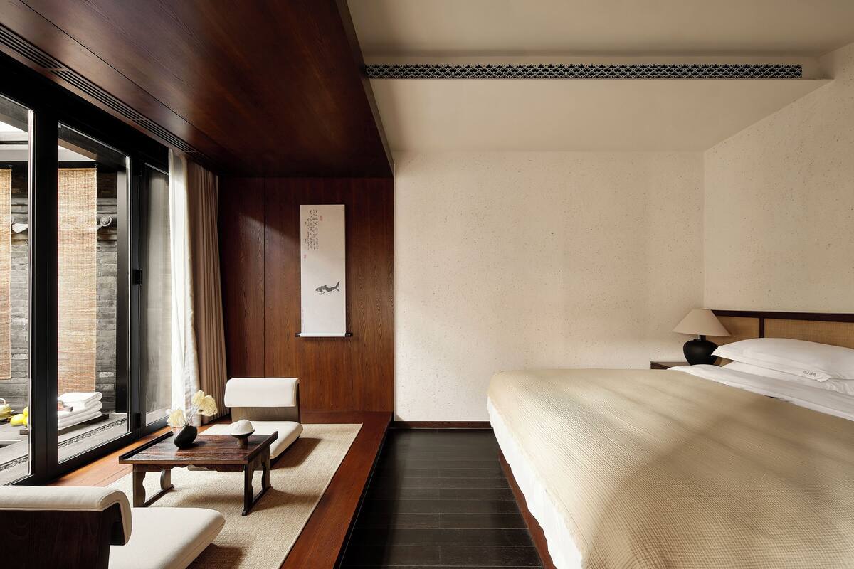 A hotel room with a bed, a small table with a vase, a chair, a nightstand with a lamp, and a view of the outside through a large window.