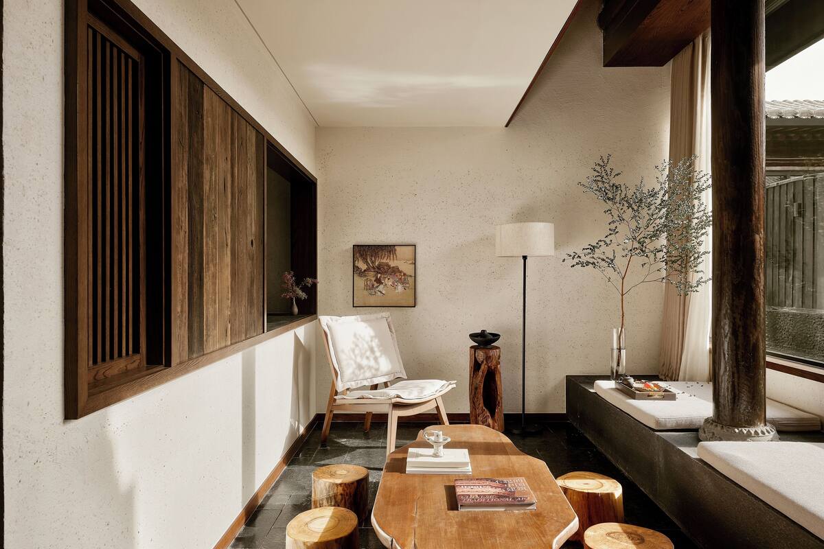 A room with a wooden table, a chair, a floor lamp, a window with a view, and a painting on the wall.