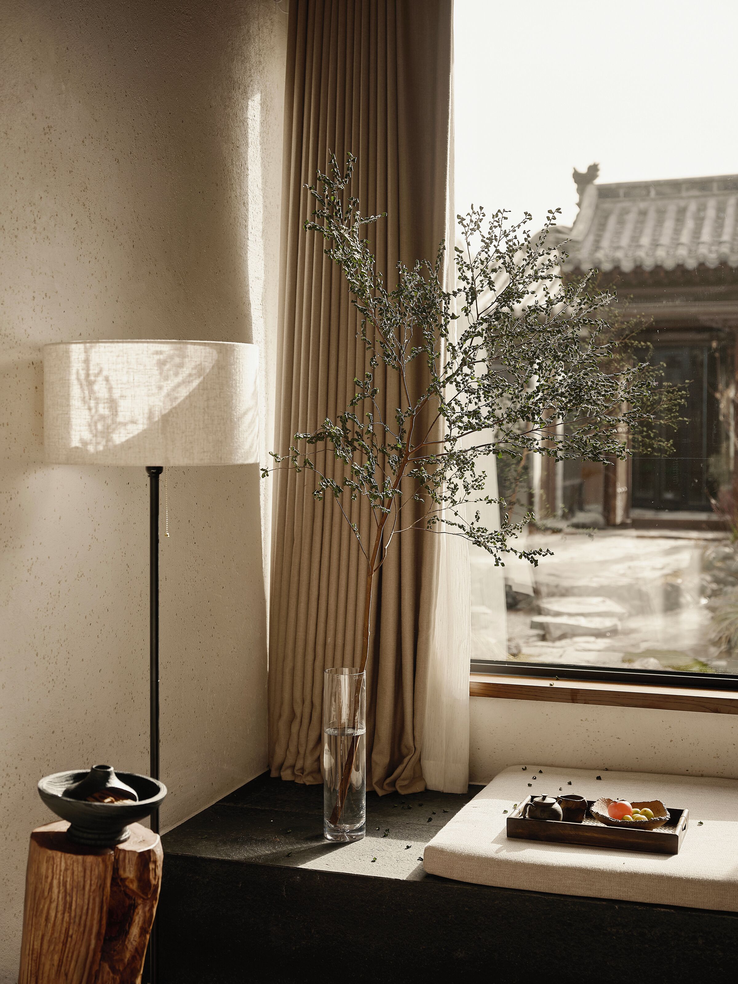 Een uitzicht op traditionele architectuur vanuit een raam, een vaas met takken, een tafel met een dienblad eten, en een lamp.
