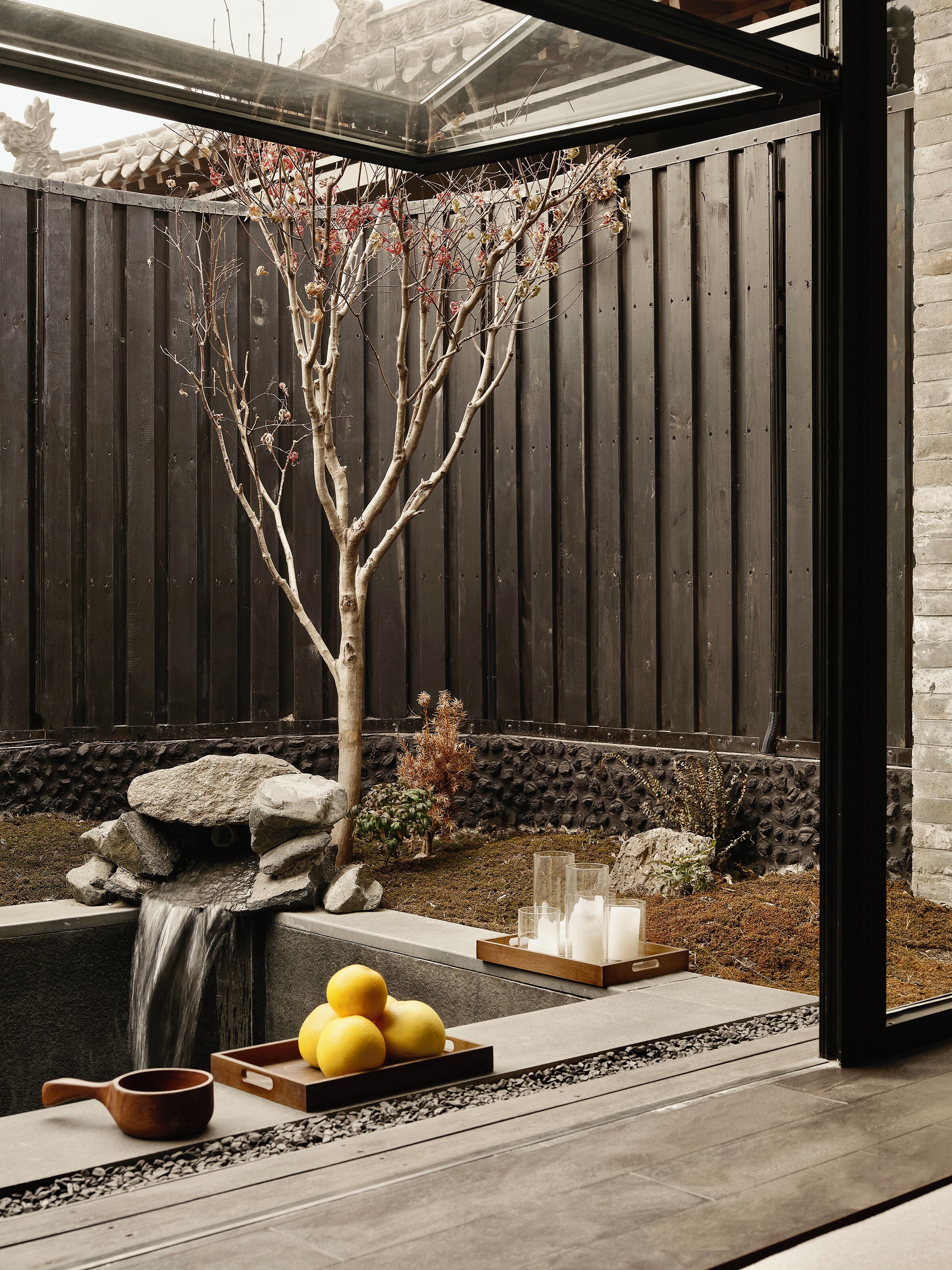A serene garden with a tree, a small waterfall, and a stone basin with lemons.
