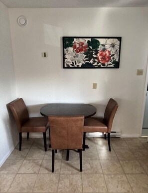 A dining area with a round table and four brown chairs, a floral painting on the wall, and a tiled floor. - Serene and Comfortable Suburban Hideaway (Lower Sackville)