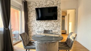 Ein moderner Essbereich mit einem runden Tisch, zwei Stühlen, einem flachbildfernseher an der Wand und Blick ins Freie durch ein großes Fenster.