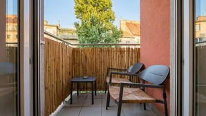 Ein Balkon mit Holzlattenzaun, zwei Stühlen, einem kleinen Tisch und Blick auf Gebäude und einen Baum.