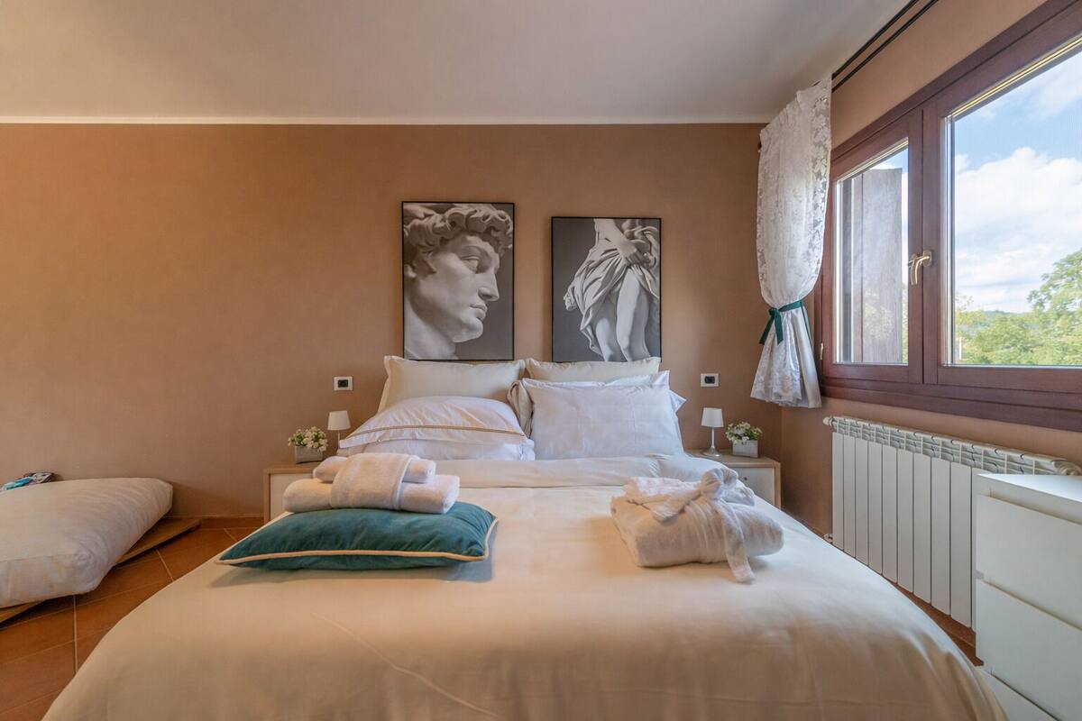 Un dormitorio con cama, almohadas, una ventana con cortinas y obras de arte en la pared.