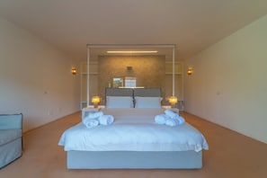 Quarto de hotel moderno com uma cama grande, duas mesas de cabeceira com abajures, vista de um edifício pela janela e uma pequena área de estar.