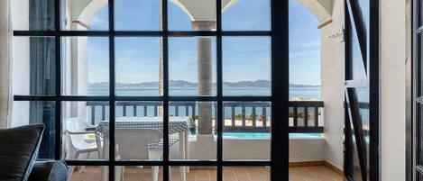 A balcony with a view of the sea, a pool, and a table with chairs.