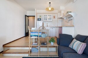 A living room with a sofa, a wooden shelf with books and decorative items, a staircase, and a kitchen area in the background. - Cargol De Mar, sea views and pool (L'Escala)