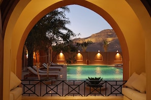 An indoor swimming pool with lounge chairs and a view of a desert landscape through an arched doorway.