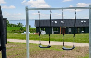 A swing set with a metal frame and two swings, a dirt path, and a building with a dark roof in the background. - Pet friendly home in Faaborg with sauna (Faaborg)