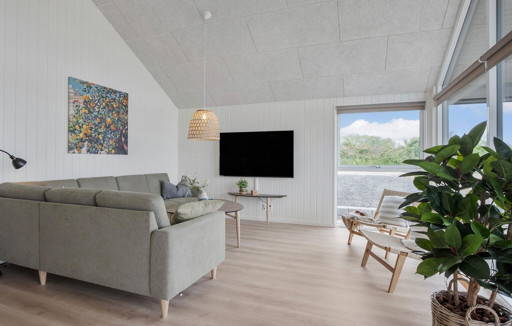 A modern living room with a grey sofa, a flat-screen TV, and a large window offering a view of greenery.