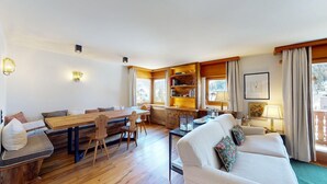 Ein gemütlicher Wohnbereich mit einer Holzlounge, einem Sofa, einem Esstisch und einem Bücherregal.