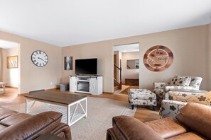 A living room with a TV, sofa, ottoman, and a clock on the wall.