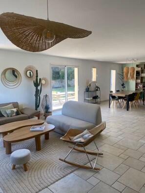A spacious living room with a large sofa, a coffee table, a swing chair, and a dining area with a table and chairs. - Sunny villa with pool - South of France (PEYNIER)