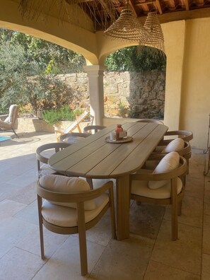 A covered outdoor dining area with a wooden table and cushioned chairs, stone walls, and hanging light fixtures. - Sunny villa with pool - South of France (PEYNIER)