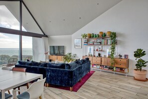 A modern living room with a large window, a dark blue sofa, a wooden cabinet with books, and a television. - 5 bedroom Modern beach house hot tub and sauna massive sea views beach access (Charmouth)