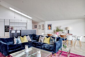 Una sala de estar moderna con un sofá de terciopelo azul, una mesa de centro de vidrio y un comedor con sillas blancas y una mesa.