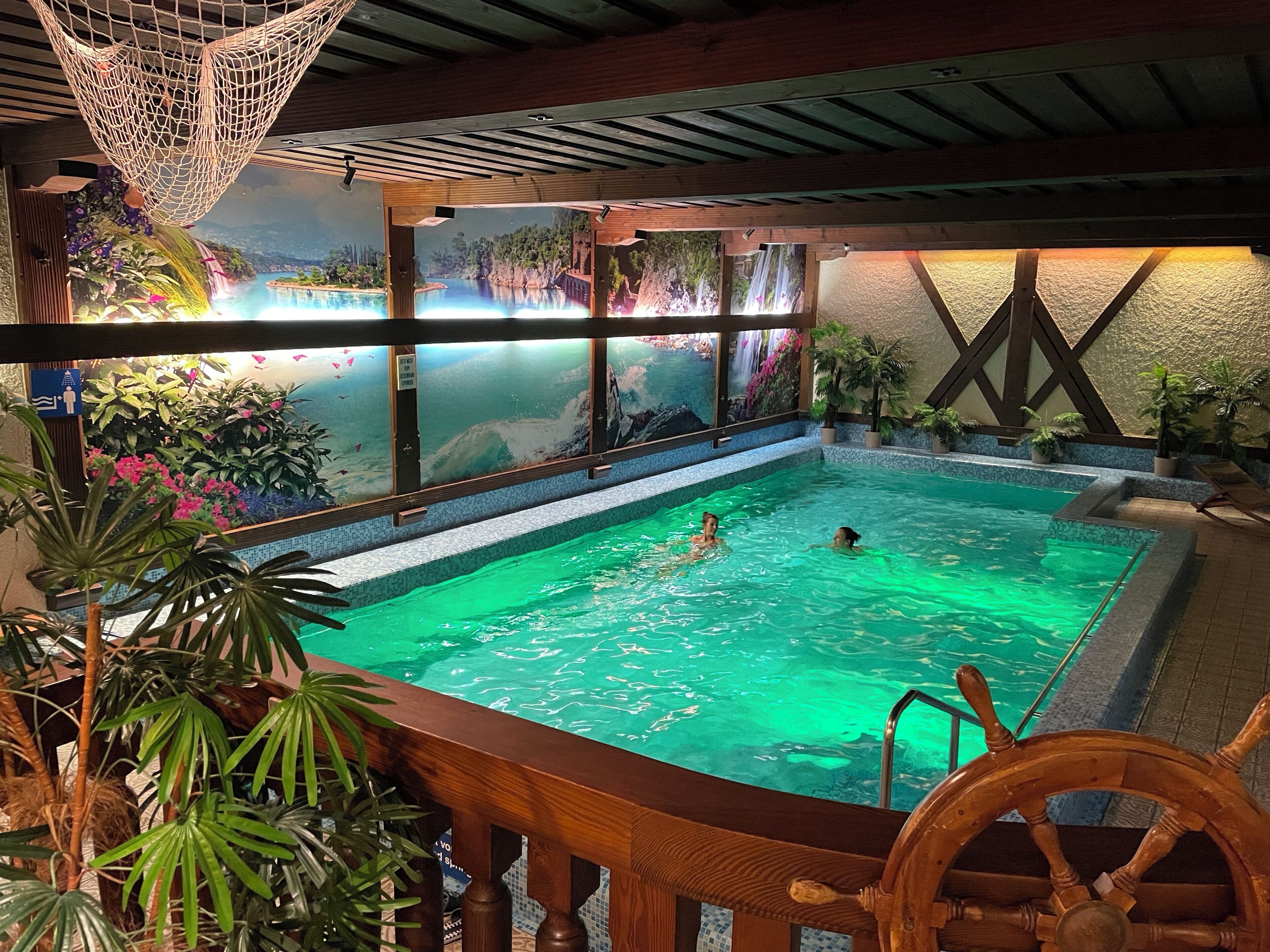 An indoor swimming pool with a wooden deck, a ship's wheel, and a mural of a tropical landscape.
