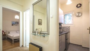 Quarto com cama, radiador, corredor que leva a um banheiro e área de cozinha com pia e relógio.