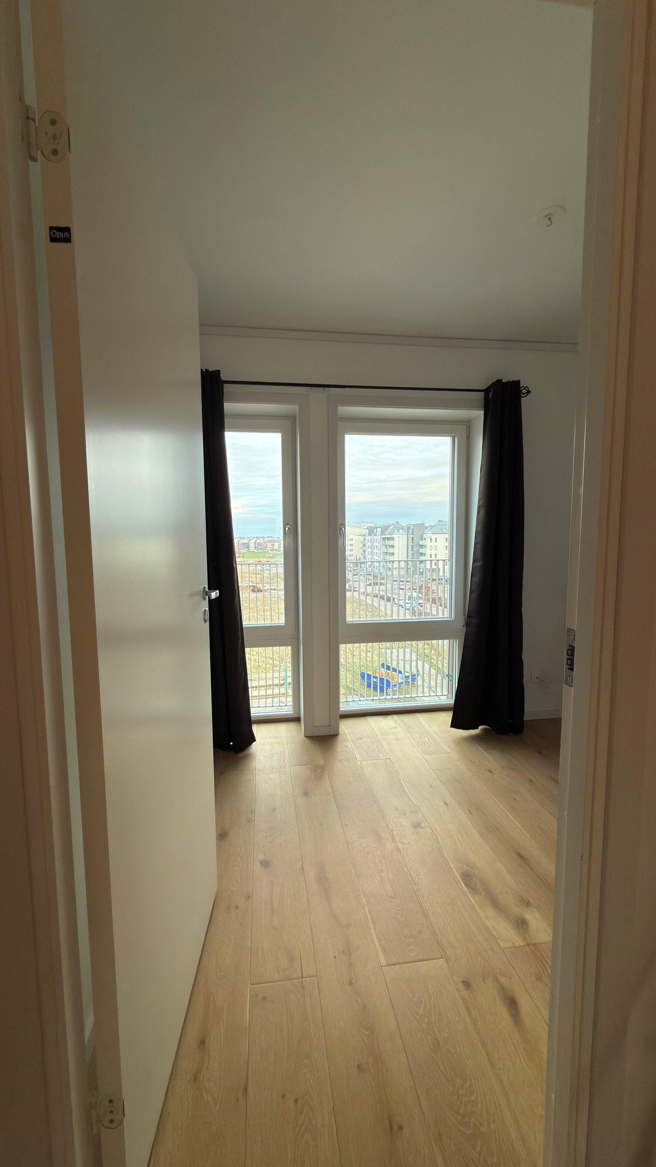 Una habitación con piso de madera, ventanas amplias y vista a edificios.