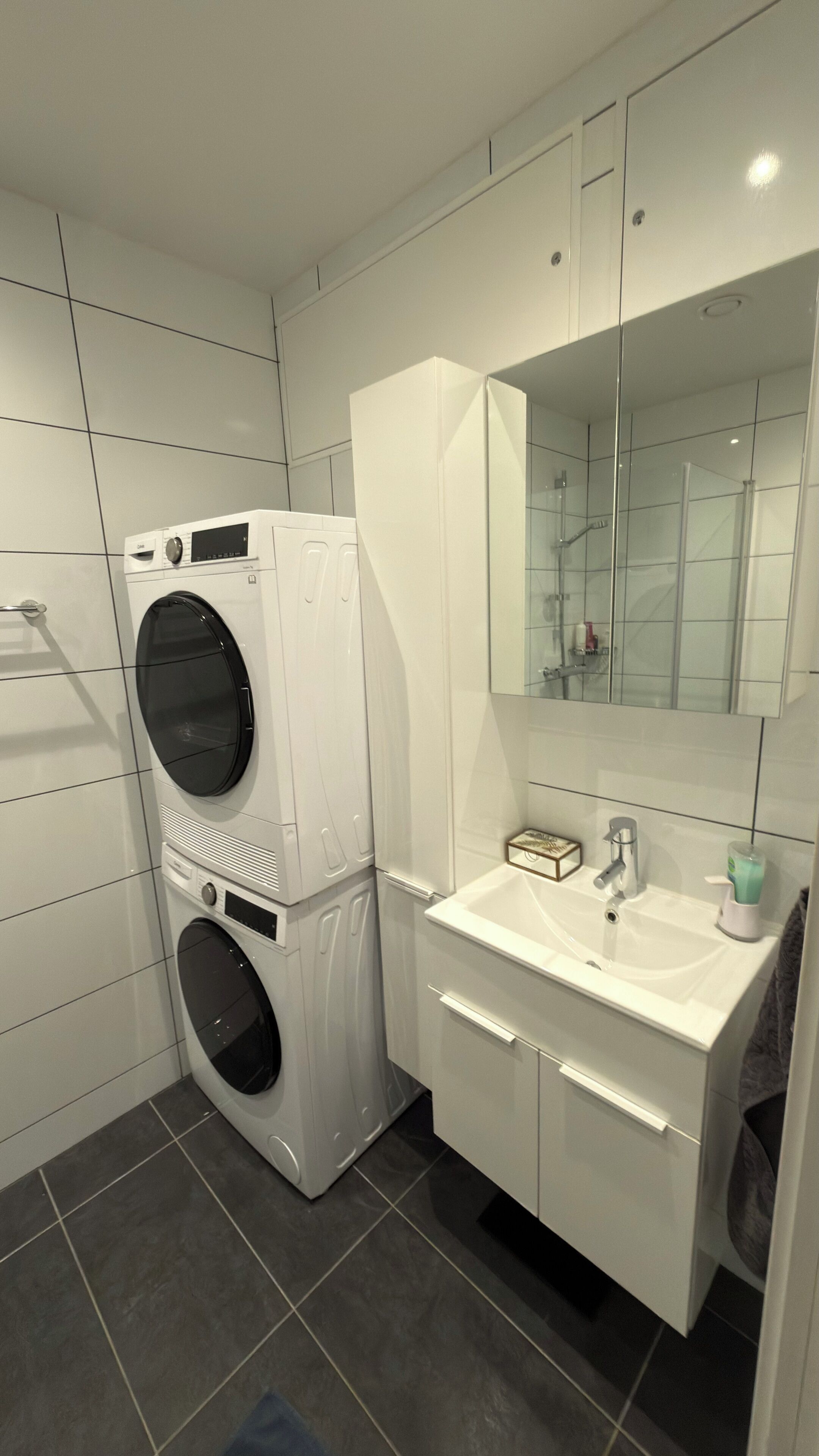Un baño moderno con lavadora y secadora apiladas, un espejo grande y un tocador blanco con lavabo.
